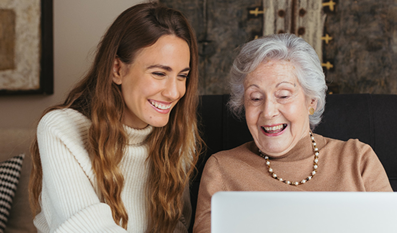 Una chica joven enseña algo en el ordenador a una anciana mientras ríen.
