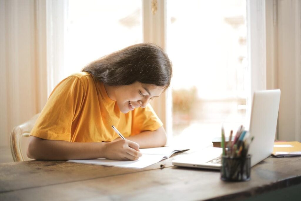 mujer joven latina con camiseta amarilla escribe sobre papel frente al ordenador