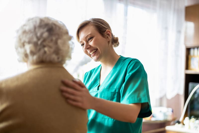 Una auxiliar de enfermería con su bata apoya a una señora mayor en un hospital. 