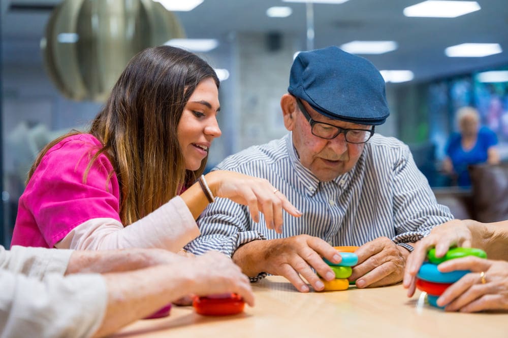 Una cuidadora ayuda a montar unos elementos de colores a un anciano en una mesa. 